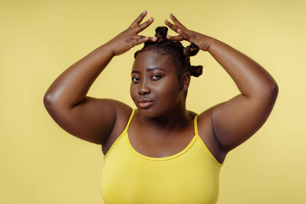Portret czarującej pięknej afrykańskiej kobiety naprawiającej stylową fryzurę patrząc na aparat odizolowany na żółtym tle. Atrakcyjna kobieta pozująca do zdjęć. Koncepcja piękna - Zdjęcie, obraz