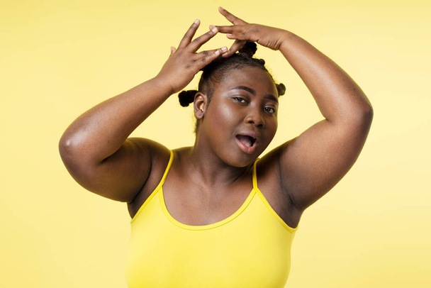 Portrait of beautiful African american woman fixing stylish hairstyle looking at camera isolated on yellow background. Attractive body positive female posing for picture. Natural beauty concept - Foto, Imagem