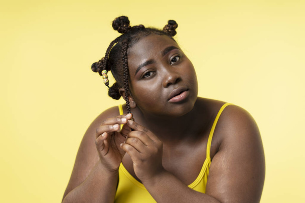 Beautiful confident Nigerian plus size woman fixing stylish hairstyle looking in mirror isolated on yellow background. Cute body positive female, natural beauty, advertisement - Фото, зображення