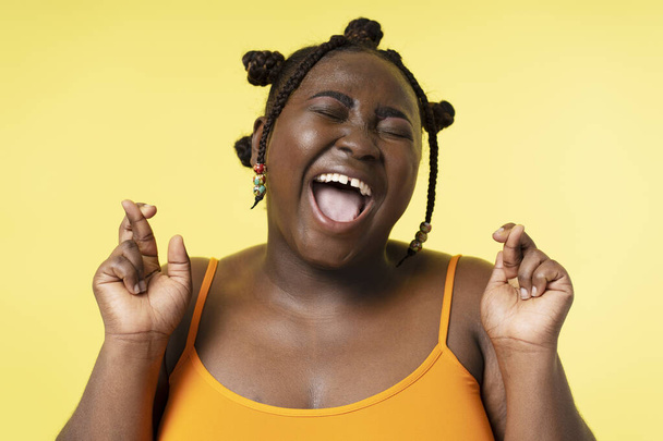 Closeup portrait of African American woman with closed eyes and crossed fingers isolated on yellow background. Smiling plus size female gesturing, hope concept - Fotó, kép