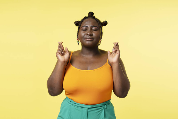 Authentic portrait of African woman with closed eyes and crossed fingers isolated on yellow background. Plus size female with stylish hairstyle gesturing, hope concept - Foto, imagen