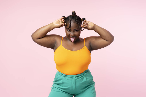 Portrait of funny African woman fooling around, looking at camera isolated on pink background. Cheerful plus size female wearing casual clothes posing for picture - Фото, изображение