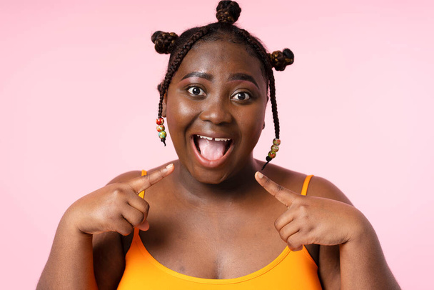 Крупный план портрета взволнованной улыбающейся женщины со стильной афро-прической, указывающей пальцами на рот, изолированный на розовом фоне. Позитивный женский жест, глядя в камеру, веселый образ жизни - Фото, изображение