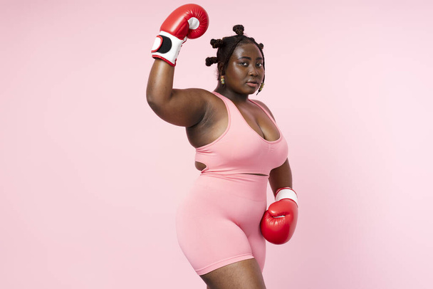 Pensive bela mulher nigeriana vestindo esportes uniforme luvas de boxe vermelho olhando para a câmera isolada no fundo rosa. Corpo autêntico modelo africano positivo posando para imagem, espaço de cópia - Foto, Imagem