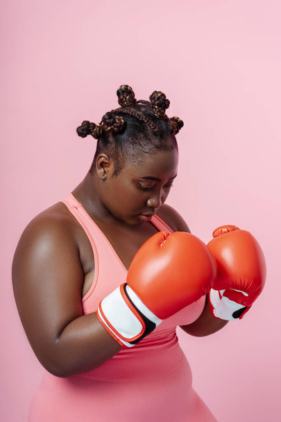 Портрет серьезной африканской женщины в красных боксёрских перчатках изолирован на розовом фоне. Обучение молодых боксеров в тренажерном зале - Фото, изображение
