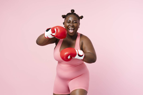 Sonriente mujer afro-americana positiva con uniforme deportivo, guantes de boxeo rojos mirando a la cámara aislada sobre fondo rosa. Nigeria entrenamiento de la deportista, atención de la salud, estilo de vida - Foto, imagen