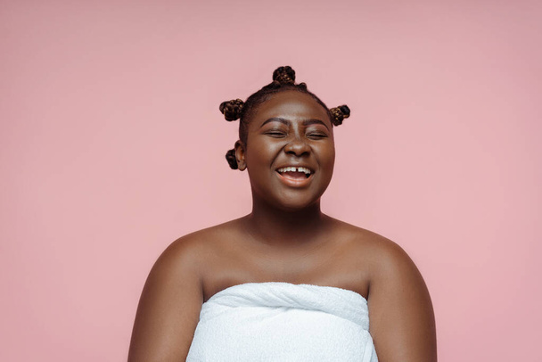 Πορτρέτο του χαμογελαστού σώματος θετική αφρικανική γυναίκα μετά το ντους σε πετσέτα απομονώνονται σε ροζ φόντο. Πρωινή ρουτίνα, φροντίδα του σώματος - Φωτογραφία, εικόνα
