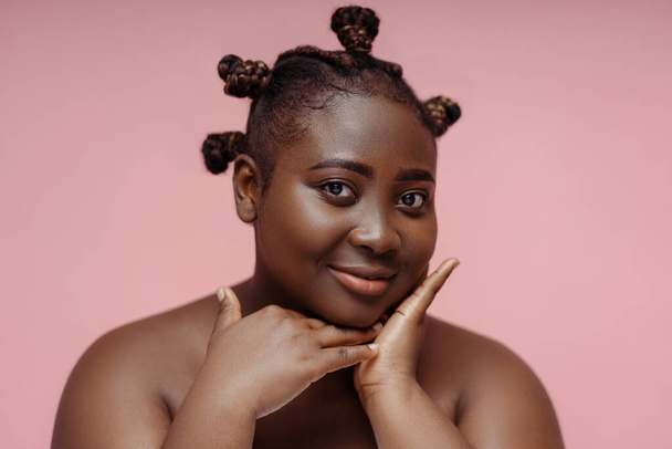 Portrait of attractive african american woman with stylish hairstyle looking in mirror touching face isolated on pink background. Beauty concept, healthy lifestyle, plus size model - Φωτογραφία, εικόνα