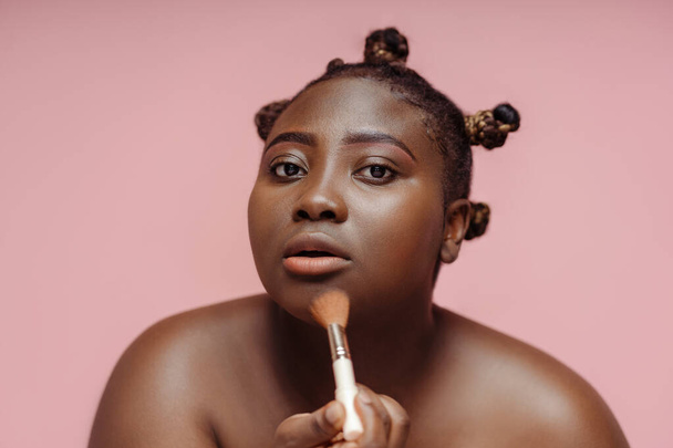 Focused African woman doing makeup, using makeup brush, looking in mirror isolated on pink background. Beauty concept, makeup, cosmetics - Foto, imagen