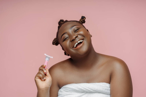 Iloinen hymyilevä afrikkalainen nainen suihkun jälkeen pyyhkeessä pitäen partaveitsi eristetty vaaleanpunainen tausta. Kauneus menettelyt, aamu rutiini, plus koko malli - Valokuva, kuva