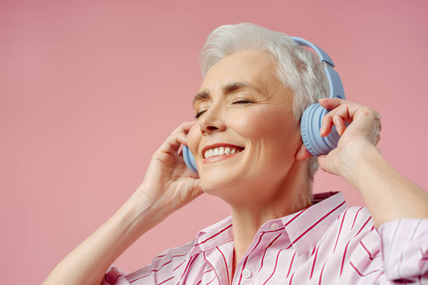 Mujer mayor feliz con los ojos cerrados escuchando música en auriculares inalámbricos, divertirse aislado ob fondo rosa. Tecnología, concepto de estilo de vida positivo  - Foto, Imagen
