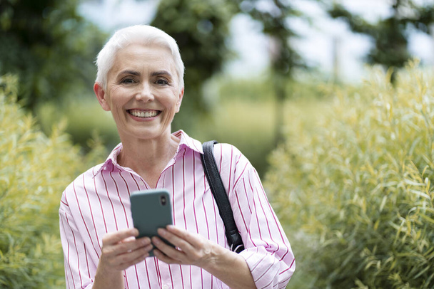 Femme mûre souriante tenant un téléphone mobile, faisant du shopping en ligne dans la rue, copiant de l'espace. Belle femme âgée aux cheveux gris vérifiant le courrier. Concept technologique - Photo, image