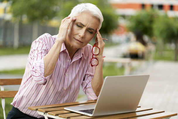 Donna anziana stanca che tiene occhiali rossi, usando il computer portatile, avendo mal di testa all'aperto. Triste donna d'affari seduta sul posto di lavoro. Problemi di salute, emicranie. - Foto, immagini
