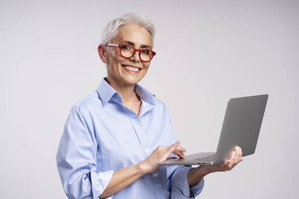 Portret uśmiechniętej pięknej dojrzałej kobiety noszącej stylowe czerwone okulary, korzystającej z laptopa, patrzącej na kamerę odizolowaną na szarym tle. Siwe włosy bizneswoman, udana koncepcja biznesu - Zdjęcie, obraz