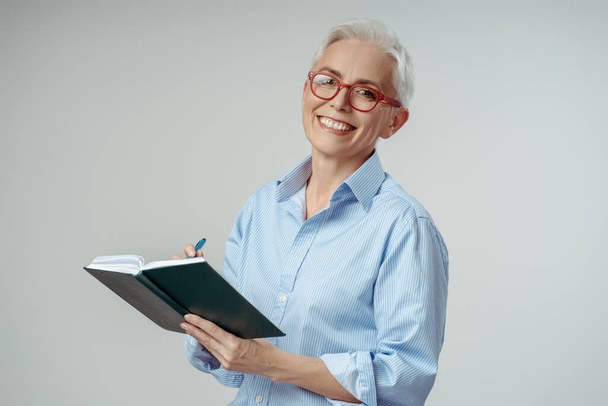 Atrakcyjna siwowłosa kobieta w stylowych okularach, czytająca książkę, planująca projekt odizolowany na szarym tle. Starszy student, nauka języka, przestrzeń do kopiowania. Koncepcja edukacji - Zdjęcie, obraz