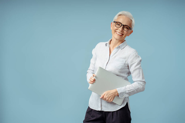 Уверенный улыбающийся менеджер, деловая женщина с ноутбуком, смотрящая в камеру на синем фоне. Счастливая поза седого фрилансера. Успешная бизнес-концепция  - Фото, изображение