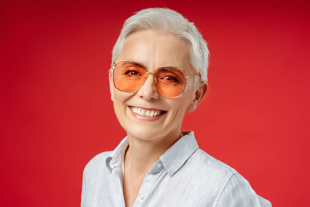 Портрет усміхненої сіро-волосистої старшої жінки в стильних сонцезахисних окулярах ізольовано на червоному тлі. Приваблива бізнес-леді, генеральний директор, менеджер, дивлячись на камеру. Успішний бізнес  - Фото, зображення