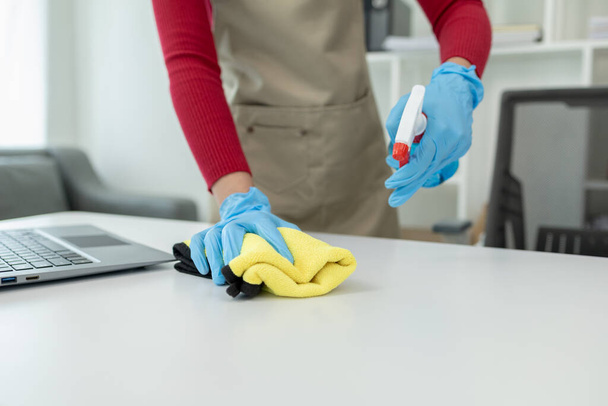 Ofisteki masa yüzeyini dezenfektan spreyiyle temizlemek, eldiven giymek ve masayı havluyla silmek, temizlikçi, temizlik için çalışma masasını temizliyor çünkü Covid-19, temizlik fikri.... - Fotoğraf, Görsel