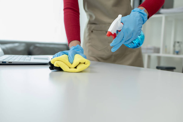 Nettoyage de la surface du bureau avec un désinfectant vaporisateur, porter des gants et essuyer la table avec une serviette, la femme de ménage nettoie le bureau pour l'hygiène en raison de la Covid-19, idée de nettoyage. - Photo, image