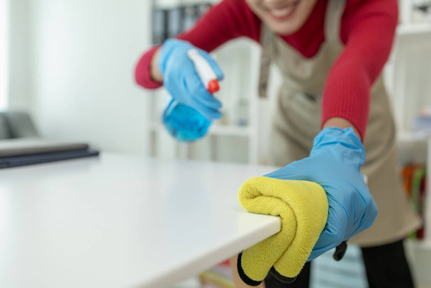 Aasialainen naispuolinen siivooja pyyhkii pöydät puhdistussuihkeella, käyttää kumikäsineitä ja esiliinaa ja työskennellä onnellisella hymyllä, Pyyhi pöytä pyyhkeellä, puhdistusaine.. - Valokuva, kuva