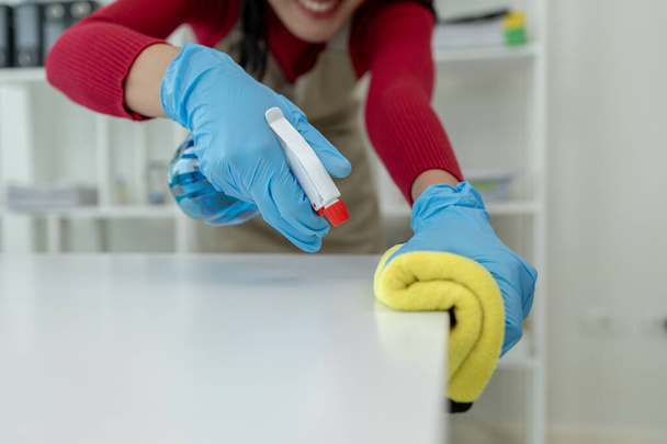 Ασιάτισσα καθαρίστρια σκουπίζει τα τραπέζια με σπρέι καθαρισμού, φορούν λαστιχένια γάντια και ποδιά και εργάζονται με ένα χαρούμενο χαμόγελο, Χρησιμοποιήστε μια πετσέτα για να σκουπίσει το τραπέζι, την ιδέα καθαρισμού. - Φωτογραφία, εικόνα