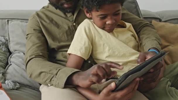 Tilt up von schwarzem Mann mit frühem Sohn beim gemeinsamen Spielen auf Tablet-PC in gemütlicher häuslicher Atmosphäre - Filmmaterial, Video