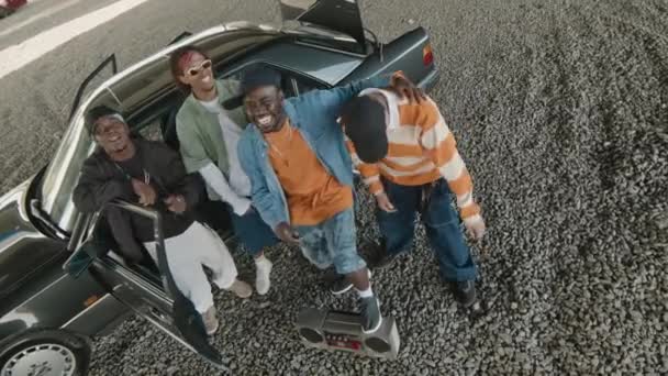 Afrika kökenli Amerikalı ve Kafkas kökenli dört kişinin eski moda müzik setlerinde siyah eski moda arabalarının yanında durup hip hop dinledikleri yüksek açılı bir çekim. - Video, Çekim