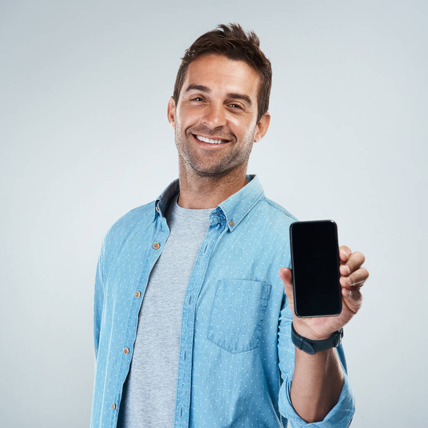Co si myslíš o mém novém telefonu? Portrét veselého mladého muže držícího a zobrazujícího svůj mobil, zatímco stojí na šedém pozadí - Fotografie, Obrázek