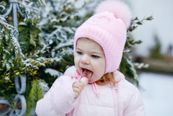 Adorable little toddler girl walking outdoors in winter. Cute toddler eating sweet lollypop candy. Child having fun on cold snow day. Wearing warm baby pink clothes and hat with bobbles - Photo, Image