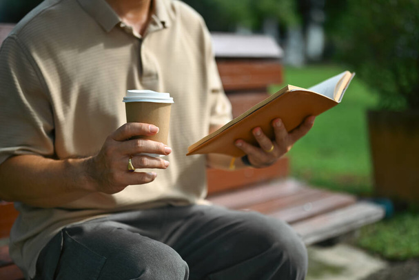 Middle aged and drinking coffee and reading book while sitting on bench at summer park. - Photo, Image