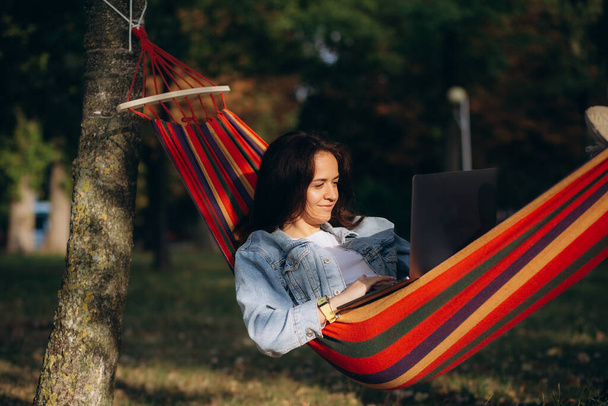 Молода красива жінка лежить в гамаку з ноутбуком в саду. зелений фон. Дерева. телефонна робота. віддалена робота. Високоякісна фотографія - Фото, зображення