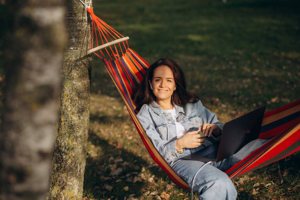 Kobieta siedząca na trawie w parku, pracująca na laptopie. Kobieta nosząca słuchawki za pomocą laptopa siedząc pod drzewem w parku z jasnym słońcem od tyłu.  - Zdjęcie, obraz