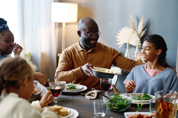 Egy fiatal, mosolygós férfi tálcát ad át sült kukoricával az egyik lányának, miközben mellette ül az ünnepi asztalnál, házi készítésű ételekkel tálalva. - Fotó, kép