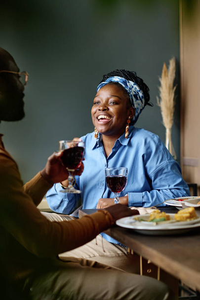 Νεαρή χαρούμενη γυναίκα κοιτάζει τον άντρα της με ένα ποτήρι κόκκινο σπιτικό κρασί να κάθεται μπροστά της σερβίροντας γιορτινό τραπέζι. - Φωτογραφία, εικόνα