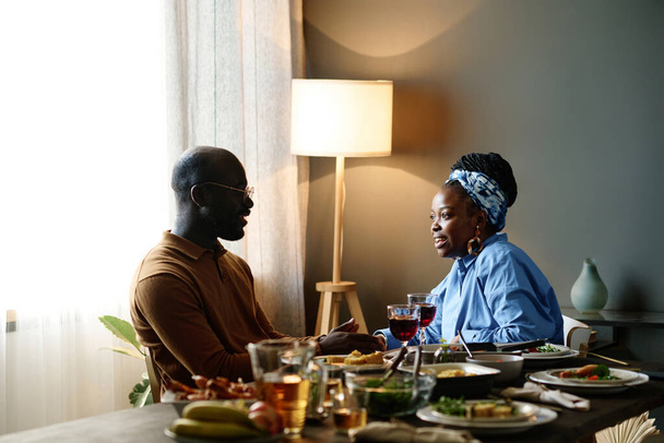 Vue latérale du jeune couple afro-américain discutant autour d'une table de fête servie avec de la nourriture et des boissons faites maison pendant le dîner à la maison - Photo, image