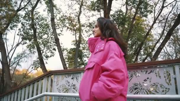 Dame élégante et souriante en veste rose marchant dans le parc de la ville d'automne tout en descendant les escaliers. Automne style urbain de mode de rue. Saison d'automne. Temps réel - Séquence, vidéo