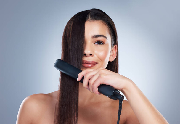 Beautiful young woman smiling while using a hair straightener on her long brown hair. Brunette woman using flat iron. Using hair care products to prevent heat damage. - Photo, Image