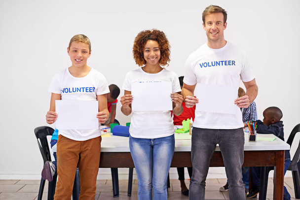 Helfen ist seine eigene Belohnung. Porträt von Freiwilligen, die ein leeres Schild hochhalten, während sie mit kleinen Kindern arbeiten - Foto, Bild
