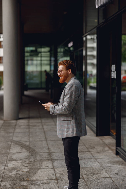 Handsome male freelancer holding phone while standing near office during break time  - Photo, Image