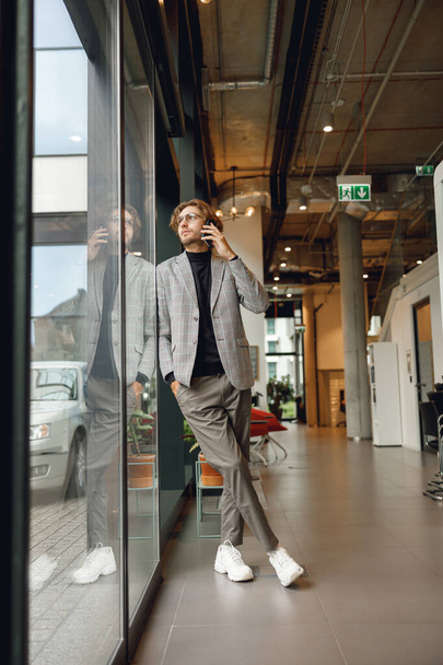 Bonito homem de negócios falando por telefone celular em pé no escritório perto da janela e olha para longe - Foto, Imagem
