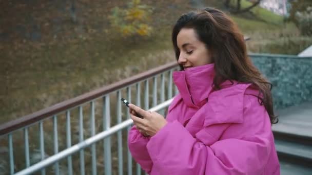Ελκυστική νεαρή κοπέλα που χρησιμοποιεί το κινητό τηλέφωνο, ενώ πληκτρολογεί το μήνυμα, περνώντας χρόνο στο πάρκο της πόλης. Φθινόπωρο αστικό στυλ μόδας δρόμο. Φθινοπωρινή. Πραγματικός χρόνος - Πλάνα, βίντεο