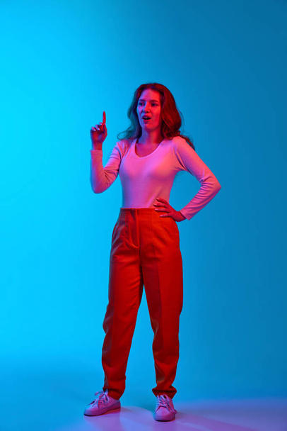 Full-length image of young girl in casual clothes raising finger up, generating ideas against blue studio background in neon light. Concept of youth, human emotions, facial expression, lifestyle - Zdjęcie, obraz