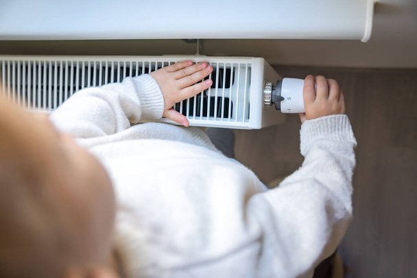 Little girl in warm sweater and socks turning the battery heating knob and warming up from the heating radiator. Heating in an apartment, at home. High quality photo - Photo, Image