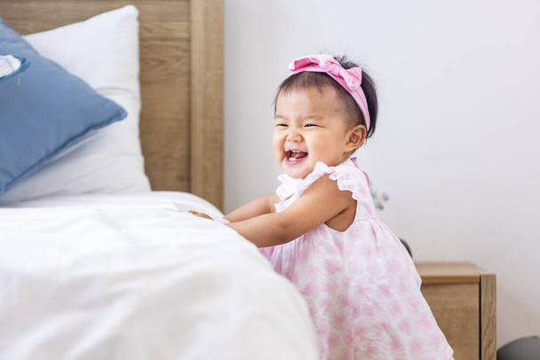Asiatisches Kleinkind in süßem rosa Kleid lächelt, während es mit dem Gehen beginnt und sich am Bett balanciert mit Glück für gesundes Kind und entzückendes Mädchenporträt - Foto, Bild