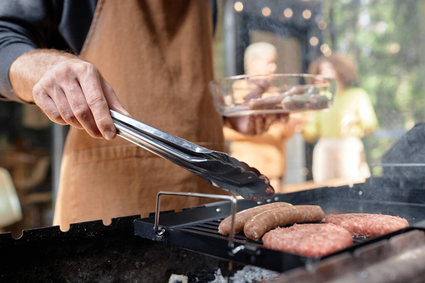 Close-up van een jonge man in een schort die worstjes maakt op de grill tijdens de picknick - Foto, afbeelding