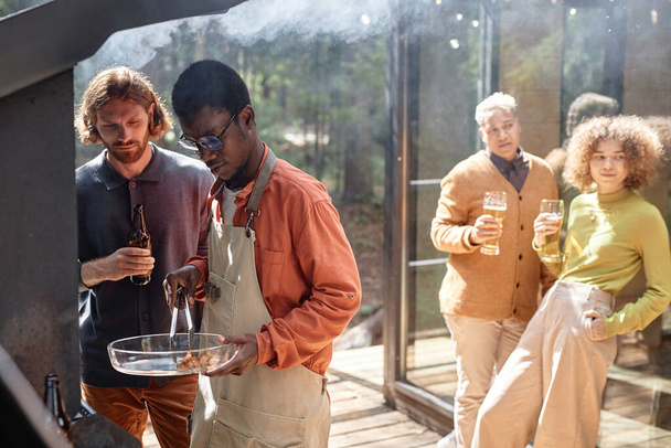 Vriendengroep die vlees koken en bier drinken tijdens de picknick in het bos - Foto, afbeelding
