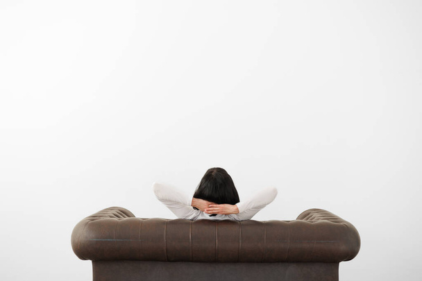 Rear view at relaxed woman with hands behind head sits on comfortable leather sofa on white wall background, feels mental balance, dreams of satisfaction, enjoys no stress free peaceful. Copy space - Photo, Image