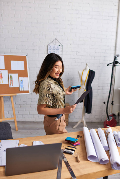 innoittamana aasialainen stylisti tarkastellaan väri swatches lähellä kannettavaa tietokonetta ja ompelu kuvioita studio, muoti - Valokuva, kuva