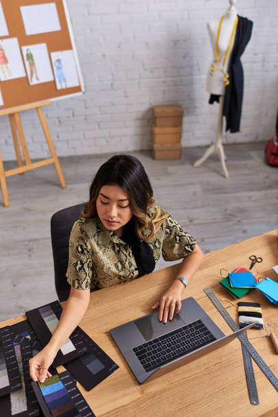 kreativní asijský módní návrhář při pohledu na barevné vzorníky v blízkosti notebooku na pracovním stole, vysoký úhel pohledu - Fotografie, Obrázek