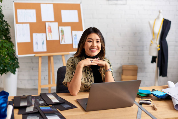 веселый азиатский стилист смотрит в камеру рядом с ноутбуком и цветные образцы на рабочем столе в ателье - Фото, изображение